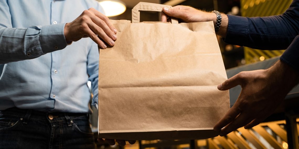 Man in shirt picking a takeaway food package from a restaurant. Blank craft paper box with room for text or logo. Crop shot, unrecognizable faces.