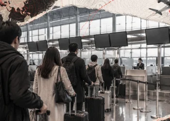 Terminal aeroportuale: viaggiatori di spalle con valigie in coda ai banchi bag drop, banconi e luce diurna.